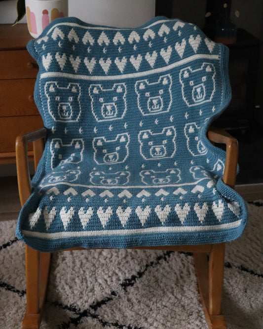 Blue knitted blanket with bear pattern draped over a wooden chair on a carpeted floor.